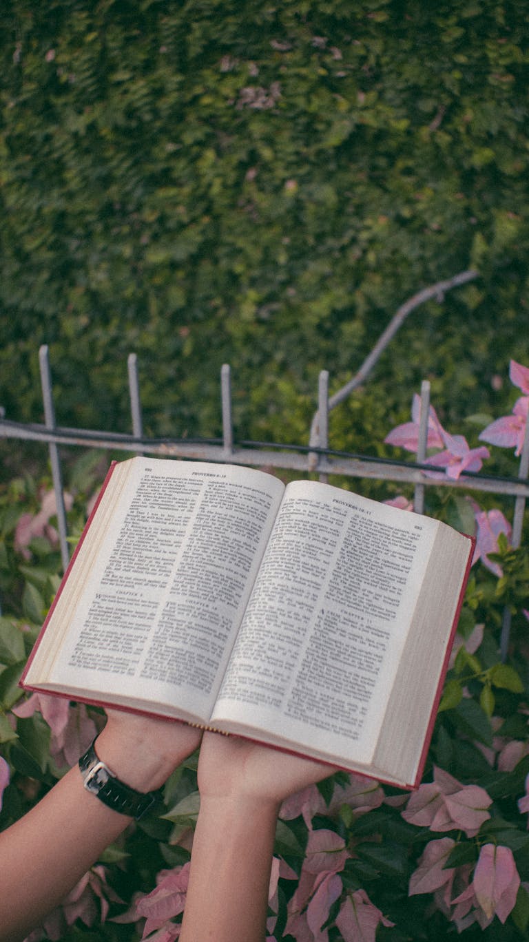 Open book held by hands amidst pink flowers and green garden background.